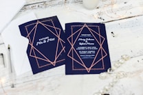 Wedding invitations are placed on a rustic white wooden table. The invitations feature a navy blue background with elegant geometric designs in rose gold. One card displays the words 'Future Mr & Mrs,' while the other provides details about the wedding, including names and the date. Nearby are a pen, an envelope, and some string lights, creating a warm and inviting atmosphere.