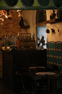 A vintage-style interior decorated with dark wooden furniture, an old radio, and a vintage fan. The walls have a rustic design with brickwork and antique light fixtures. Shelves hold various decorative vases and teapots, adding to the classic ambiance.