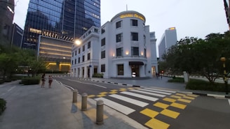 A modern urban street scene captures a grand white building with the name 'Maxwell Chambers' prominently displayed. Tall glass office towers and lush greenery surround the area. A patterned pedestrian crossing with yellow and white stripes is in the foreground. Street lighting adds to the ambiance.