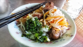 A bowl of Asian noodle salad featuring grilled meat, fresh green onions, shredded carrots, and white radish. The dish is garnished with crushed peanuts and accompanied by a pair of black chopsticks resting on the bowl. A glass of water is visible next to the bowl on a reflective surface.