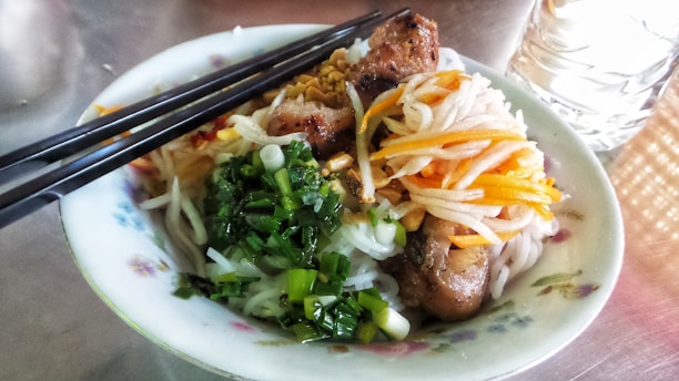 A bowl of Asian noodle salad featuring grilled meat, fresh green onions, shredded carrots, and white radish. The dish is garnished with crushed peanuts and accompanied by a pair of black chopsticks resting on the bowl. A glass of water is visible next to the bowl on a reflective surface.