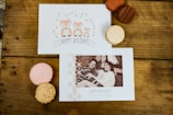 Holiday cards and macarons are arranged on a wooden table. One card features an illustration of foxes and the text 'Happy Holidays' while the other has a family portrait with a holiday greeting. The macarons come in various colors, including pink, orange, and brown.