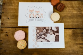 Holiday cards and macarons are arranged on a wooden table. One card features an illustration of foxes and the text 'Happy Holidays' while the other has a family portrait with a holiday greeting. The macarons come in various colors, including pink, orange, and brown.