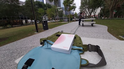 A park setting with a pathway flanked by grass and trees. A book titled 'Winning with Honour' is resting on a backpack placed on a bench. In the background, a person is sitting at a picnic table.