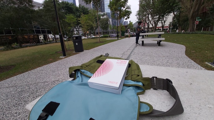 A park setting with a pathway flanked by grass and trees. A book titled 'Winning with Honour' is resting on a backpack placed on a bench. In the background, a person is sitting at a picnic table.