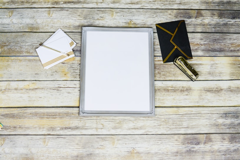 A flat lay of a stationary set featuring a stack of blank white paper placed in the center, surrounded by a pen with a polka-dot pattern, a few white envelopes, a black envelope with gold trim, and a metallic gold stapler all arranged on a rustic wooden surface.