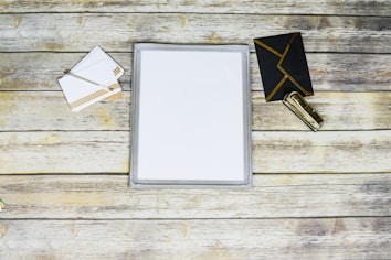 A flat lay of a stationary set featuring a stack of blank white paper placed in the center, surrounded by a pen with a polka-dot pattern, a few white envelopes, a black envelope with gold trim, and a metallic gold stapler all arranged on a rustic wooden surface.