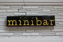 A neon sign with the word 'minibar' illuminated in yellow. The sign is mounted on a white painted brick wall, with visible black electrical wiring above it.