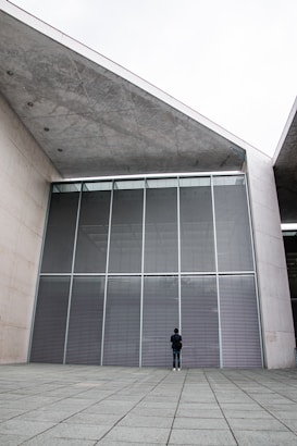 A person stands alone facing a large building with modern architecture. The structure features expansive metal and glass paneling, creating a reflective surface. The concrete surroundings give a minimalist and austere vibe, enhancing the feeling of scale and solitude.