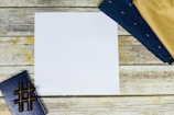 A blank white sheet of paper is centered on a wooden surface, surrounded by a closed blue notebook with a hashtag symbol and folded dark blue and beige fabric pieces.