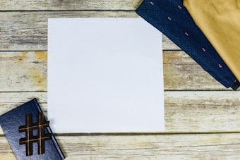 A blank white sheet of paper is centered on a wooden surface, surrounded by a closed blue notebook with a hashtag symbol and folded dark blue and beige fabric pieces.