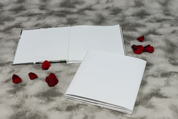 An open and a closed booklet are placed on a textured gray surface. Scattered around the booklets are several red rose petals, adding a touch of color to the composition.
