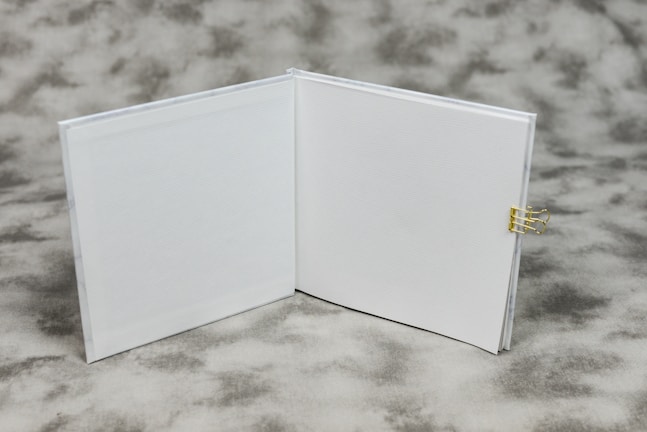 An open book with blank white pages is displayed on a textured gray background. A small gold binder clip is attached to the right side of the book's cover.