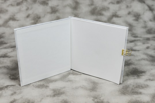 An open book with blank white pages is displayed on a textured gray background. A small gold binder clip is attached to the right side of the book's cover.