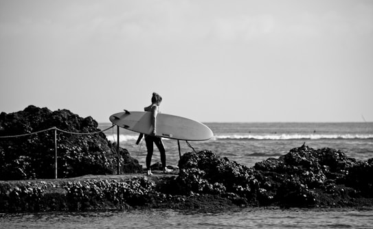 A person is standing on a rocky shore, holding a surfboard under one arm, and looking towards the ocean. The waves in the background create a serene and open atmosphere, while the black and white filter adds a classic and timeless feel.