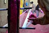 A person is engaging in a handcraft process, applying vibrant pink dye to threads stretched across a wooden frame, possibly for textile making. The background features additional people blurred in motion, suggesting a busy workshop environment.