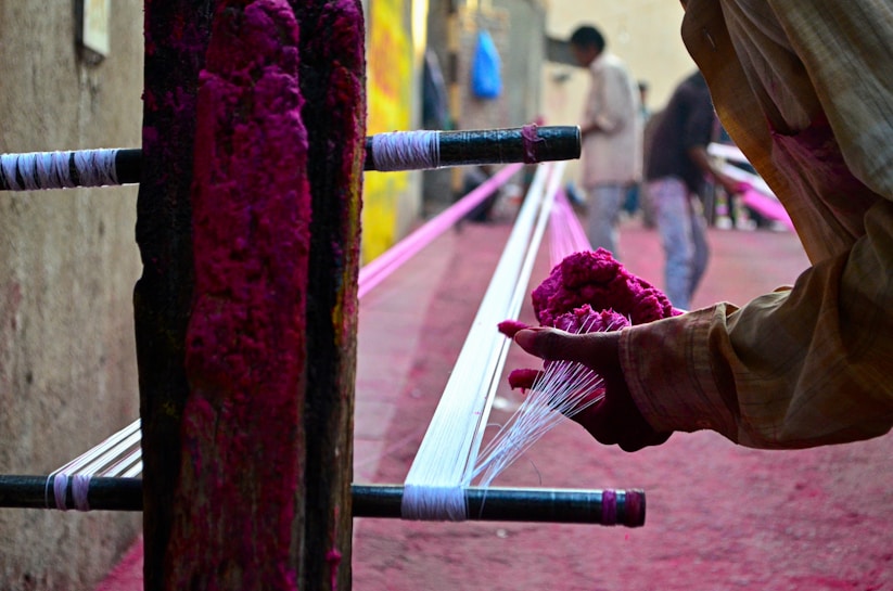 A person is engaging in a handcraft process, applying vibrant pink dye to threads stretched across a wooden frame, possibly for textile making. The background features additional people blurred in motion, suggesting a busy workshop environment.