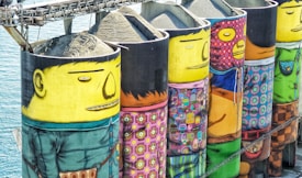 A series of large industrial silos are painted with vibrant, colorful murals depicting abstract human figures wearing various patterned clothing. The murals are set against a backdrop of water, creating a striking contrast between the industrial and artistic elements.