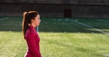 A woman in a red athletic top stands on a grass sports field with markings, seemingly focused and calm. The field appears to be part of a stadium with empty bleachers in the background.
