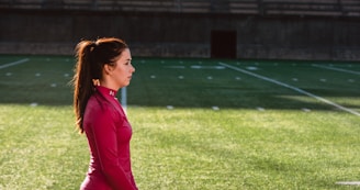 A woman in a red athletic top stands on a grass sports field with markings, seemingly focused and calm. The field appears to be part of a stadium with empty bleachers in the background.