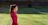 A woman in a red athletic top stands on a grass sports field with markings, seemingly focused and calm. The field appears to be part of a stadium with empty bleachers in the background.