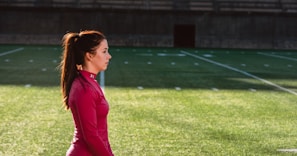 A woman in a red athletic top stands on a grass sports field with markings, seemingly focused and calm. The field appears to be part of a stadium with empty bleachers in the background.