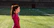 A woman in a red athletic top stands on a grass sports field with markings, seemingly focused and calm. The field appears to be part of a stadium with empty bleachers in the background.