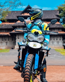 A person in a motorcycle outfit and helmet is sitting on an off-road motorbike. The motorbike features bold blue and yellow designs. In the background, there is a traditional building with a distinctly sloped roof, surrounded by some greenery under a bright blue sky.