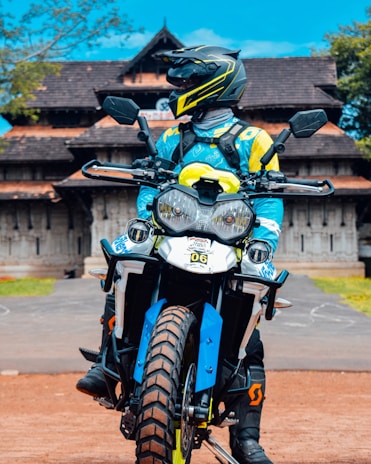 A person in a motorcycle outfit and helmet is sitting on an off-road motorbike. The motorbike features bold blue and yellow designs. In the background, there is a traditional building with a distinctly sloped roof, surrounded by some greenery under a bright blue sky.