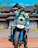 A person in a motorcycle outfit and helmet is sitting on an off-road motorbike. The motorbike features bold blue and yellow designs. In the background, there is a traditional building with a distinctly sloped roof, surrounded by some greenery under a bright blue sky.