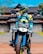 A person in a motorcycle outfit and helmet is sitting on an off-road motorbike. The motorbike features bold blue and yellow designs. In the background, there is a traditional building with a distinctly sloped roof, surrounded by some greenery under a bright blue sky.
