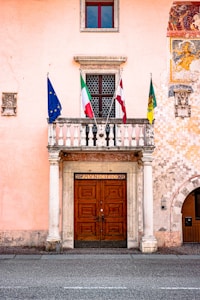 A peach-colored building facade with a large, ornate wooden double door framed by white columns. Above the door is a stone balcony with three flags: the European Union, Italy, and another unidentified flag. A decorative mural is partially visible on the right side, and a small window with a red frame is positioned above the balcony.