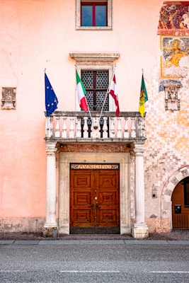 A peach-colored building facade with a large, ornate wooden double door framed by white columns. Above the door is a stone balcony with three flags: the European Union, Italy, and another unidentified flag. A decorative mural is partially visible on the right side, and a small window with a red frame is positioned above the balcony.