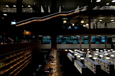A modern library interior with two levels. The lower level features rows of tables where individuals are studying under soft, warm lighting. There are bookshelves filled with books on both levels. The upper level includes walkways and additional shelving, with a few people visible in silhouette.