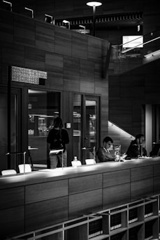 A monochromatic indoor scene featuring a consultation area with a modern design. Two people are seated at a long wooden table, working on laptops, while another person stands nearby. Bookshelves are visible beneath the table, and a sign reads 'Consultation Specialisee'. Overhead lighting and glass partitions add to the contemporary architectural style.