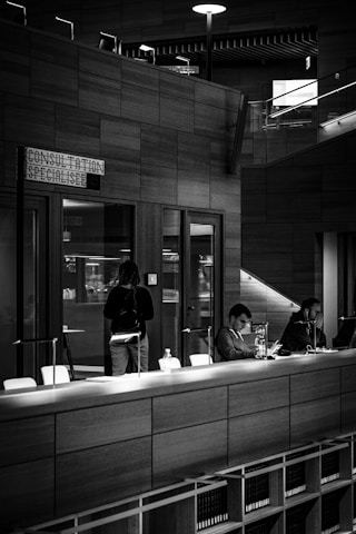 A monochromatic indoor scene featuring a consultation area with a modern design. Two people are seated at a long wooden table, working on laptops, while another person stands nearby. Bookshelves are visible beneath the table, and a sign reads 'Consultation Specialisee'. Overhead lighting and glass partitions add to the contemporary architectural style.