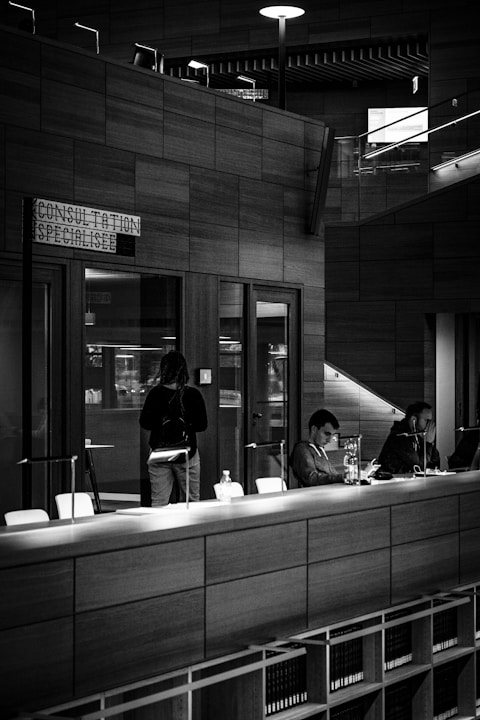 A monochromatic indoor scene featuring a consultation area with a modern design. Two people are seated at a long wooden table, working on laptops, while another person stands nearby. Bookshelves are visible beneath the table, and a sign reads 'Consultation Specialisee'. Overhead lighting and glass partitions add to the contemporary architectural style.
