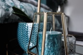 A teal-colored candle is enclosed in a geometric glass holder with gold accents. Behind it, there are decorative ceramic pots, one with a herringbone pattern and another with intricate gray details. A green plant leaf is partially visible, adding a touch of nature to the scene.