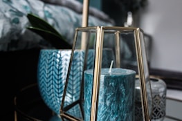 A teal-colored candle is enclosed in a geometric glass holder with gold accents. Behind it, there are decorative ceramic pots, one with a herringbone pattern and another with intricate gray details. A green plant leaf is partially visible, adding a touch of nature to the scene.