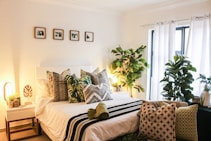 A cozy bedroom with a neatly made bed topped with an assortment of decorative pillows, featuring green and botanical patterns. The room is decorated with lush indoor plants, adding a natural touch. A small side table with a plant and lamp is next to the bed. Light filters through sheer white curtains, creating a warm and inviting ambiance. Several small botanical prints are hung on the wall above the bed.