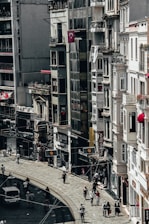 A busy city street lined with tall buildings. Numerous people are walking along the sidewalk, and a few vehicles can be seen on the road. The architecture appears urban and densely packed, with various shop signs and Turkish flags displayed.