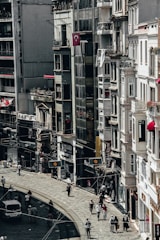 A busy city street lined with tall buildings. Numerous people are walking along the sidewalk, and a few vehicles can be seen on the road. The architecture appears urban and densely packed, with various shop signs and Turkish flags displayed.