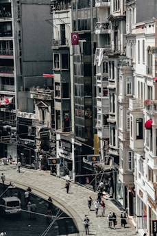 A busy city street lined with tall buildings. Numerous people are walking along the sidewalk, and a few vehicles can be seen on the road. The architecture appears urban and densely packed, with various shop signs and Turkish flags displayed.