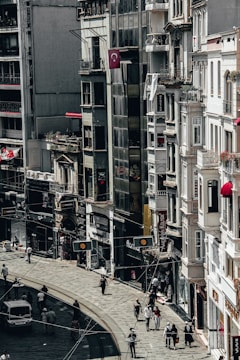 A busy city street lined with tall buildings. Numerous people are walking along the sidewalk, and a few vehicles can be seen on the road. The architecture appears urban and densely packed, with various shop signs and Turkish flags displayed.
