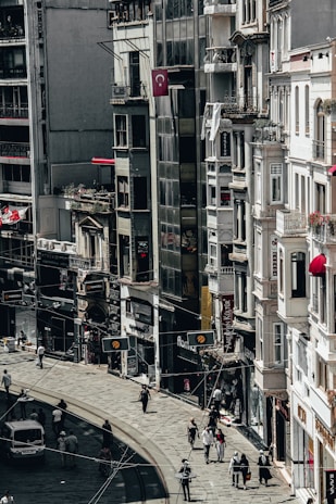A busy city street lined with tall buildings. Numerous people are walking along the sidewalk, and a few vehicles can be seen on the road. The architecture appears urban and densely packed, with various shop signs and Turkish flags displayed.
