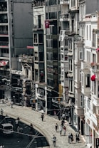 A busy city street lined with tall buildings. Numerous people are walking along the sidewalk, and a few vehicles can be seen on the road. The architecture appears urban and densely packed, with various shop signs and Turkish flags displayed.