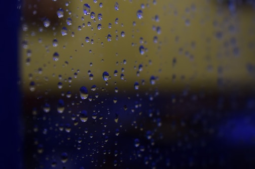 Water droplets are scattered across a glass surface, with a blurred background featuring dark blue and muted yellow hues.