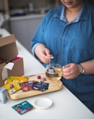 A person is preparing tea by dipping a tea bag into a clear glass cup. The person is wearing a blue denim shirt and a watch. Various colorful tea packets are spread on a wooden tray, along with a few dried rosebuds.