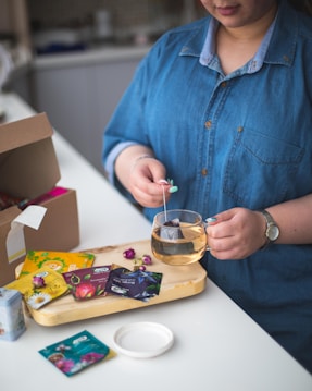 A person is preparing tea by dipping a tea bag into a clear glass cup. The person is wearing a blue denim shirt and a watch. Various colorful tea packets are spread on a wooden tray, along with a few dried rosebuds.