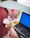 A person with a headscarf is on the phone at a desk. The workspace includes a laptop with a blue screen, a pink bottle, a cardboard box, and a packet of snacks. The person is facing away, focusing on the phone call.